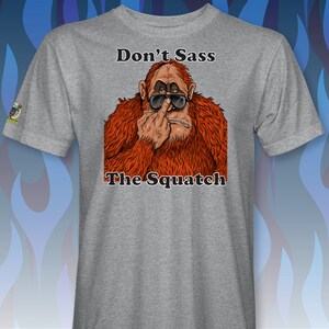 Peut inclure: Un t-shirt gris avec un graphique d'un sasquatch portant des lunettes de soleil et faisant un doigt d'honneur. Le texte "Don't Sass The Squatch" est imprim&eacute; sous l'image.