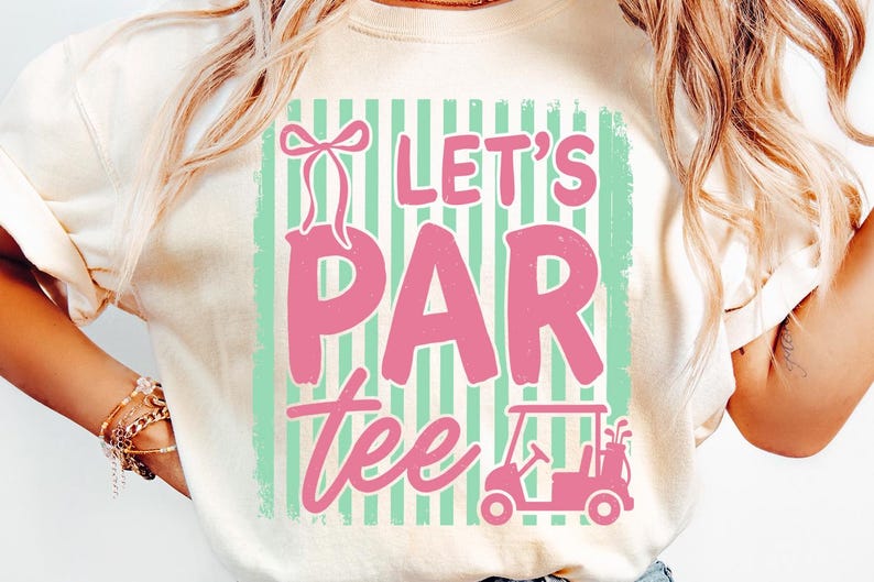 May include: A cream-coloured t-shirt featuring a distressed pink graphic that reads "Let's Par Tee". The design includes a golf cart and a bow. The background has vertical mint green and white stripes.