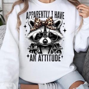 May include: White sweatshirt featuring a black and white raccoon wearing sunglasses and a leopard print bow. The text "Apparently I have an attitude" is printed on the sweatshirt.