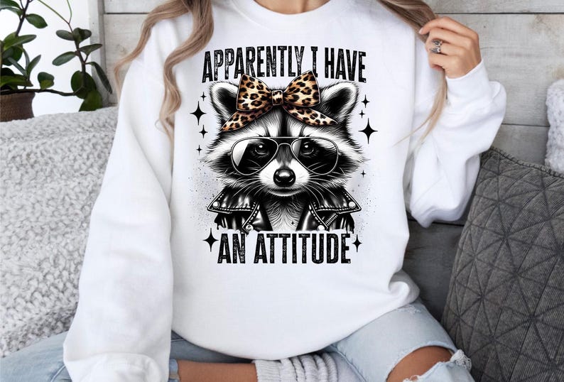 May include: White sweatshirt featuring a black and white raccoon wearing sunglasses and a leopard print bow. The text "Apparently I have an attitude" is printed on the sweatshirt.