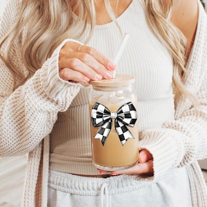 May include: A clear glass mason jar with a black and white checkered bow on the lid. The jar is filled with a light brown liquid and has a glass straw sticking out of the top.
