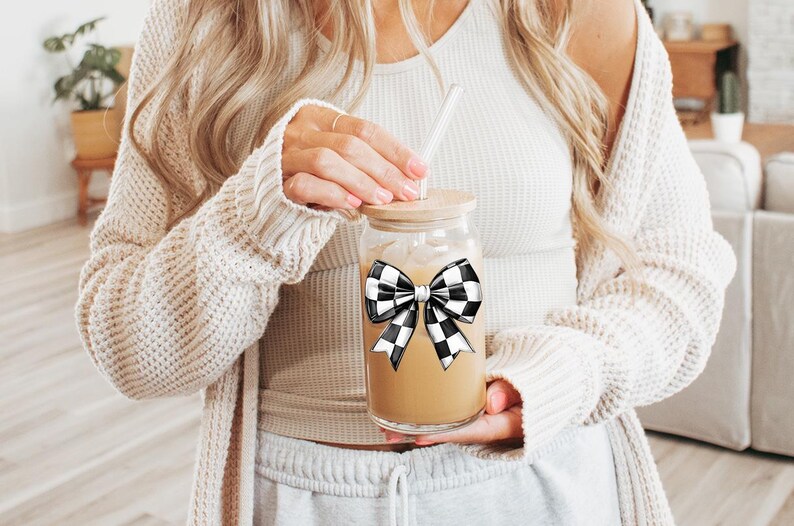 May include: A clear glass mason jar with a black and white checkered bow on the lid. The jar is filled with a light brown liquid and has a glass straw sticking out of the top.
