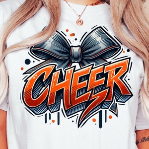 May include: White t-shirt with a graphic design of a large orange "CHEER" word with a black and gray bow on top. The design has a 3D effect and is outlined in black.