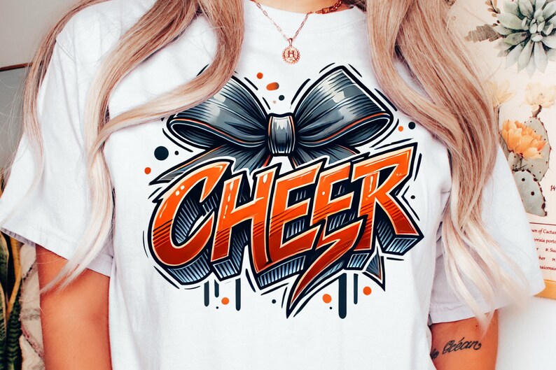 May include: White t-shirt with a graphic design of a large orange "CHEER" word with a black and gray bow on top. The design has a 3D effect and is outlined in black.