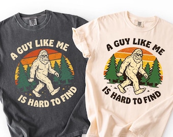 Pode incluir: Duas camisetas, uma cinza escuro e outra creme, cada uma com um gráfico de Bigfoot. O design apresenta um Bigfoot andando com óculos de sol, contra um pôr do sol e pinheiros, com o texto "A GUY LIKE ME IS HARD TO FIND."