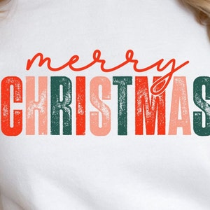Può includere: Felpa bianca con una grafica rovinata in rosso, verde e rosa che dice "buon natale" in un font scritto a mano.