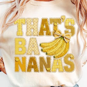May include: Cream-colored t-shirt with the phrase "THAT'S BA NANAS" in yellow patchwork letters. A bunch of bananas and a bow are also featured. The letters and bananas have a decorative trim.
