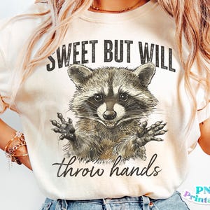 May include: A white t-shirt with a distressed graphic of a raccoon with the text "Sweet but will throw hands".
