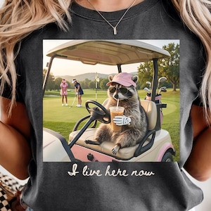 Può includere: T-shirt grigio scuro con un procione in una golf cart, con occhiali da sole e un cappello rosa, che tiene in mano una bevanda. L'immagine include il testo "I live here now" in corsivo. Sono visibili mazze da golf.