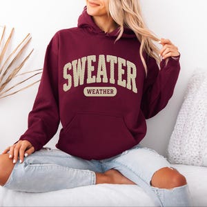May include: A maroon hoodie with the text "SWEATER WEATHER" in a cream-coloured, collegiate-style font. The hoodie features a front pocket and a drawstring hood. The model is wearing light blue jeans.
