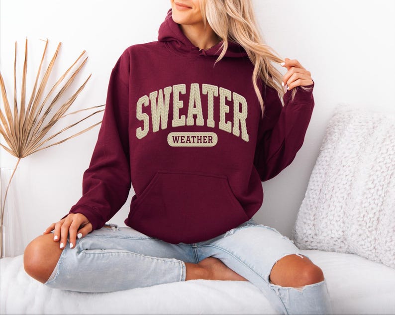 May include: A maroon hoodie with the text "SWEATER WEATHER" in a cream-coloured, collegiate-style font. The hoodie features a front pocket and a drawstring hood. The model is wearing light blue jeans.