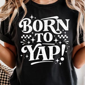 Op de afbeelding: Zwart T-shirt met de witte tekst "BORN TO YAP!" in een vetgedrukte, gestileerde lettertype. Het ontwerp bevat ster- en dambordaccenten. Een casual kledingstuk.