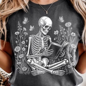 Puede incluir: Camiseta gris oscuro con un esqueleto blanco leyendo un libro, rodeado de flores blancas y mariposas. El diseño combina elementos góticos y florales, ideal para los amantes de la estética alternativa. Una camiseta gráfica única.