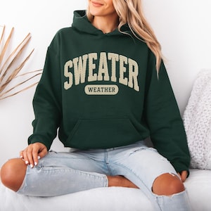 May include: A dark green hoodie with the words "SWEATER WEATHER" in a cream-coloured, collegiate-style font. The hoodie has a front pocket and a hood. The person is wearing light blue jeans.