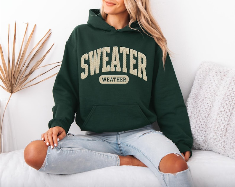 May include: A dark green hoodie with the words "SWEATER WEATHER" in a cream-coloured, collegiate-style font. The hoodie has a front pocket and a hood. The person is wearing light blue jeans.