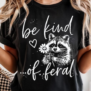 May include: Black t-shirt with the words "be kind ...of feral" in white script. A white heart and a black and white raccoon holding a daisy are also featured on the shirt.