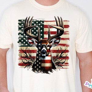May include: Cream-colored t-shirt featuring a deer head design with an American flag motif. The deer's antlers are prominent, and the flag design incorporates red and white stripes with a field of stars. The shirt is a standard crew neck.
