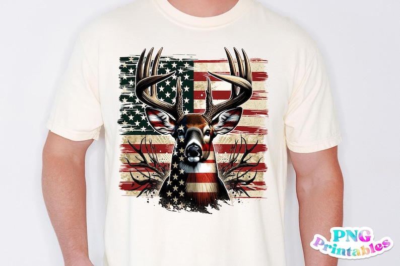 May include: Cream-colored t-shirt featuring a deer head design with an American flag motif. The deer's antlers are prominent, and the flag design incorporates red and white stripes with a field of stars. The shirt is a standard crew neck.
