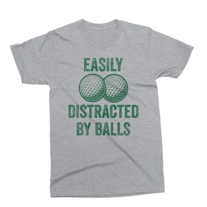 Puede incluir: Camiseta gris jaspeado con el texto verde "EASILY DISTRACTED BY BALLS". El dise&ntilde;o incluye dos pelotas de golf verdes. Cuello redondo y mangas cortas.