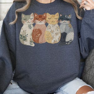Pu&ograve; includere: Felpa grigio scuro con un design applicato di quattro gatti stilizzati. Ogni gatto ha un motivo e un colore unici, tra cui beige con foglie verdi, marrone con accenti arancioni, giallo con dettagli floreali e grigio con una coda nera.