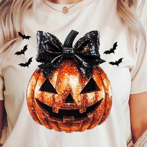 Könnte beinhalten: Ein weißes T-Shirt mit einem glitzernden orangefarbenen Kürbis mit einer schwarzen Schleife und Fledermaus-Silhouetten.