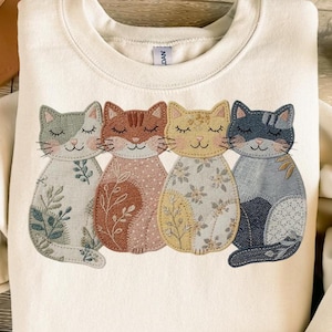 Könnte beinhalten: Cremefarbenes Sweatshirt mit einem gestickten Design von vier stilisierten Katzen. Jede Katze hat geschlossene Augen und einzigartige gemusterte Details in Grau-, Braun-, Gelb- und Blautönen. Die Katzen sind in einer Reihe angeordnet und schaffen eine skurrile und künstlerische Ästhetik.