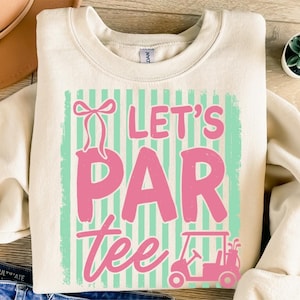 May include: A cream-coloured sweatshirt featuring the text "Let's Par Tee" in pink, set against a mint green and white striped background. A pink golf cart graphic is at the bottom. A golf-themed apparel item.