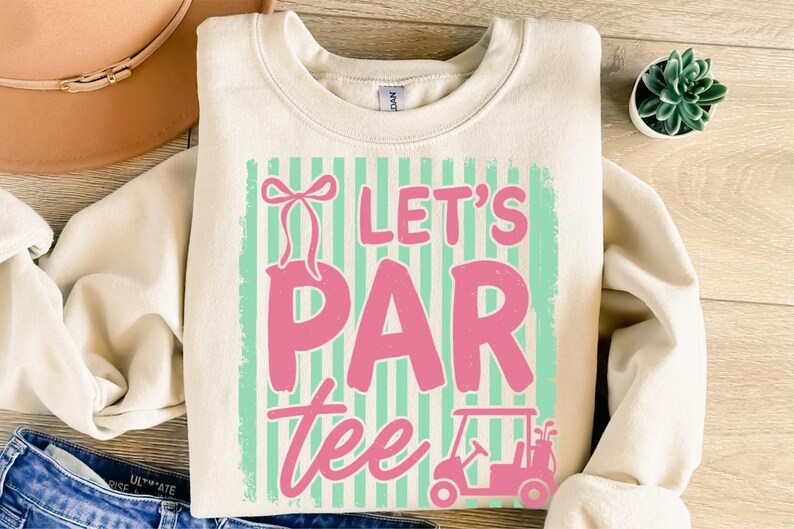May include: A cream-coloured sweatshirt featuring the text "Let's Par Tee" in pink, set against a mint green and white striped background. A pink golf cart graphic is at the bottom. A golf-themed apparel item.