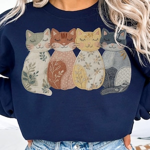 Pu&ograve; includere: Felpa blu navy con quattro applicazioni di gatti stilizzati. Ogni gatto ha un motivo unico, tra cui disegni floreali e di foglie, nei toni del crema, marrone, giallo e grigio. I gatti sono disposti in fila, con gli occhi chiusi, creando un'estetica accogliente e stravagante.