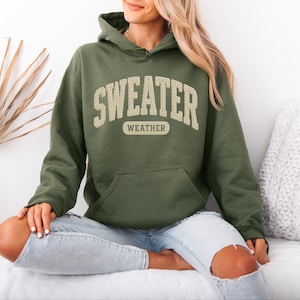 May include: An olive green hoodie with the text "SWEATER WEATHER" in a cream-coloured, vintage-style font. The hoodie has a front pocket and a drawstring hood. The model is wearing light blue jeans.
