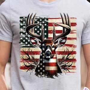 May include: A gray t-shirt featuring a graphic of a deer with antlers, superimposed over a distressed American flag design. The deer's face and chest are integrated with the flag's stars and stripes. The design is detailed and patriotic.