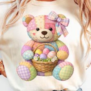 May include: A cream-colored t-shirt featuring a colorful patchwork teddy bear holding a basket of Easter eggs. The bear has a pink bow and is made of pastel-colored fabric patches. The basket contains several decorated eggs.