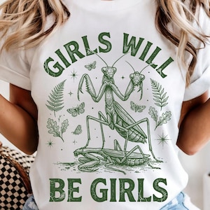 May include: White t-shirt with a green graphic design featuring a praying mantis, butterflies, and foliage. The text "GIRLS WILL BE GIRLS" is printed above and below the insect illustration. The shirt is made of a soft material.