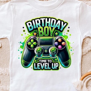May include: White t-shirt with a green, blue, and black video game controller graphic. The text "Birthday Boy" is above the controller and "Time to Level Up" is below the controller.