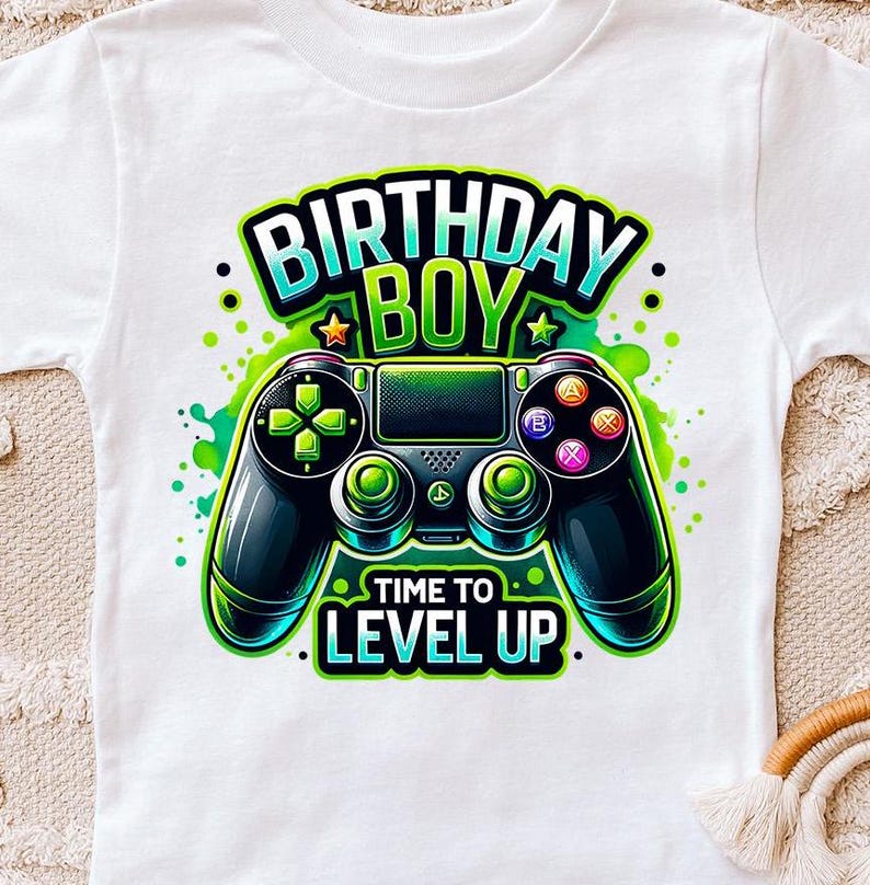 May include: White t-shirt with a green, blue, and black video game controller graphic. The text "Birthday Boy" is above the controller and "Time to Level Up" is below the controller.