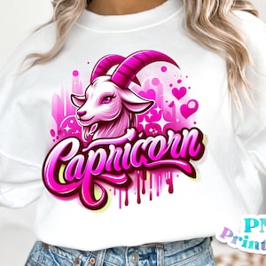 Pode incluir: Uma camisa de moletom branca com um design gr&aacute;fico rosa e branco de uma cabe&ccedil;a de cabra com a palavra "Capric&oacute;rnio" em letras rosa. O design tem um efeito aquarela rosa e branco com cora&ccedil;&otilde;es e estrelas.