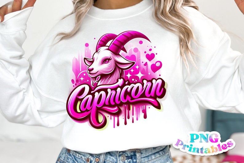 Pode incluir: Uma camisa de moletom branca com um design gr&aacute;fico rosa e branco de uma cabe&ccedil;a de cabra com a palavra "Capric&oacute;rnio" em letras rosa. O design tem um efeito aquarela rosa e branco com cora&ccedil;&otilde;es e estrelas.