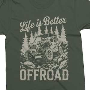 Puede incluir: Camiseta verde oscuro con un diseño gráfico. El diseño presenta un vehículo todoterreno conduciendo por un terreno rocoso, con el texto "Life is Better" arriba y "OFFROAD" abajo.