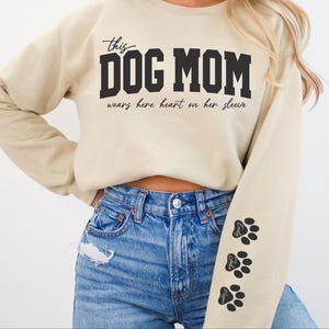 Może przedstawiać: Beżowa bluza z nadrukiem "this DOG MOM wears her heart on her sleeve" na przodzie. Bluza ma czarne odciski łap na rękawie.