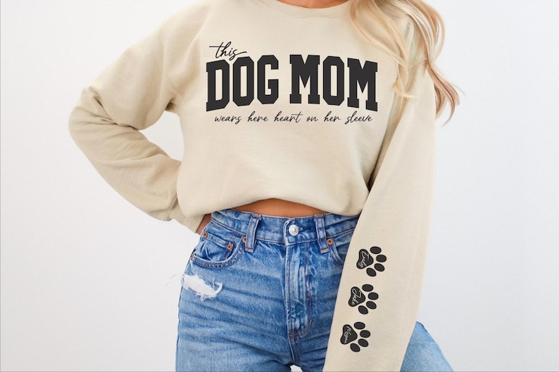 Może przedstawiać: Beżowa bluza z nadrukiem "this DOG MOM wears her heart on her sleeve" na przodzie. Bluza ma czarne odciski łap na rękawie.