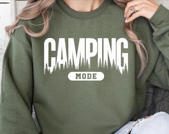 Könnte beinhalten: Ein grüner Pullover mit dem Text "CAMPING MODE" in weißen Buchstaben. Der Text ist von einer Reihe weißer Bäume umgeben.