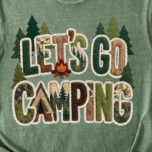 May include: Green t-shirt with the phrase "LET'S GO CAMPING" in a patchwork style. The letters are various colors and textures, including wood grain and forest patterns. Small trees and a campfire graphic are also featured.