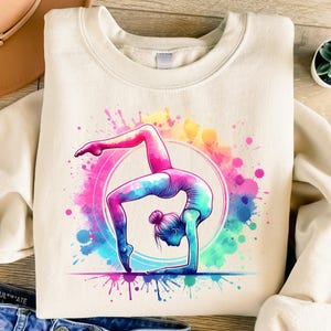 May include: A white sweatshirt with a colorful watercolor design of a woman doing a gymnastic pose. The design features pink, blue, yellow, and green watercolor splatters.