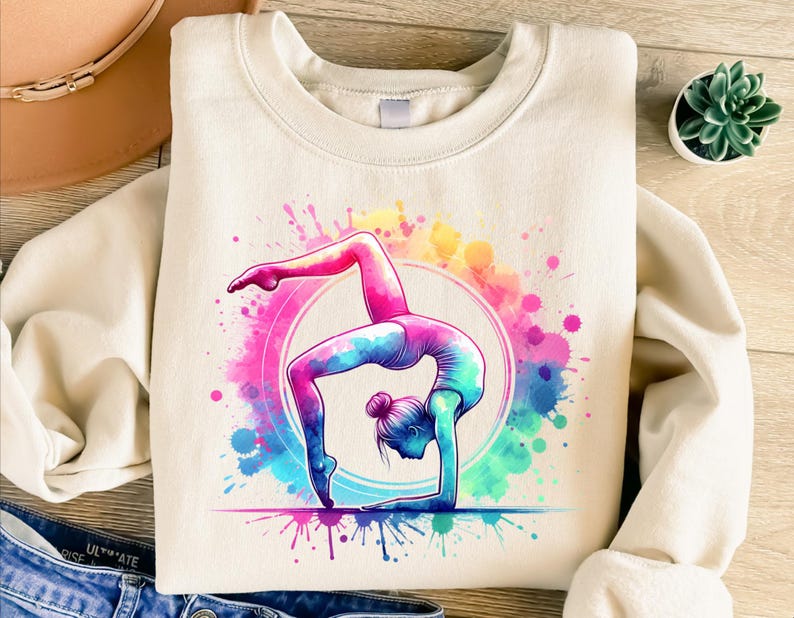 May include: A white sweatshirt with a colorful watercolor design of a woman doing a gymnastic pose. The design features pink, blue, yellow, and green watercolor splatters.