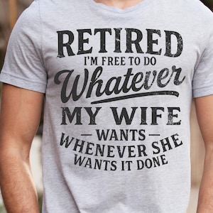 May include: A light grey t-shirt with the text "RETIRED I'M FREE TO DO WHATEVER MY WIFE WANTS WHENEVER SHE WANTS IT DONE" in black. The shirt is short-sleeved and made of a soft material.