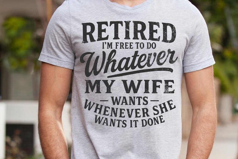 May include: A light grey t-shirt with the text "RETIRED I'M FREE TO DO WHATEVER MY WIFE WANTS WHENEVER SHE WANTS IT DONE" in black. The shirt is short-sleeved and made of a soft material.