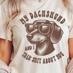 Może przedstawiać: Biała koszulka z brązowym jamnikiem w okularach przeciwsłonecznych i napisem "My Dachshund and I talk shit about you".