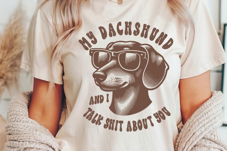 Może przedstawiać: Biała koszulka z brązowym jamnikiem w okularach przeciwsłonecznych i napisem "My Dachshund and I talk shit about you".