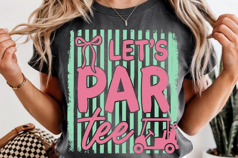 May include: Dark grey t-shirt with a pink and green graphic that says "Let's Par Tee". The design includes a golf cart and a bow. The shirt is a casual style.