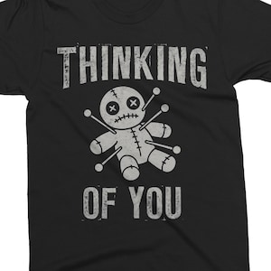 May include: Black t-shirt with the words "THINKING OF YOU" in distressed white lettering. A gray voodoo doll graphic is centered below the text, featuring cross-eyes and pins.